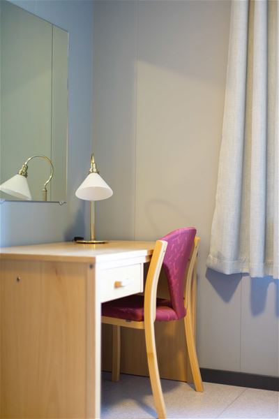 Wooden desk with a brass-and-white lamp, mirror, and a purple chair beside a window with light curtains..
