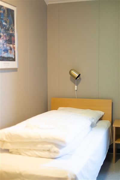 A neatly made single bed with white bedding sits below a small wall lamp, beside a nightstand and framed art..