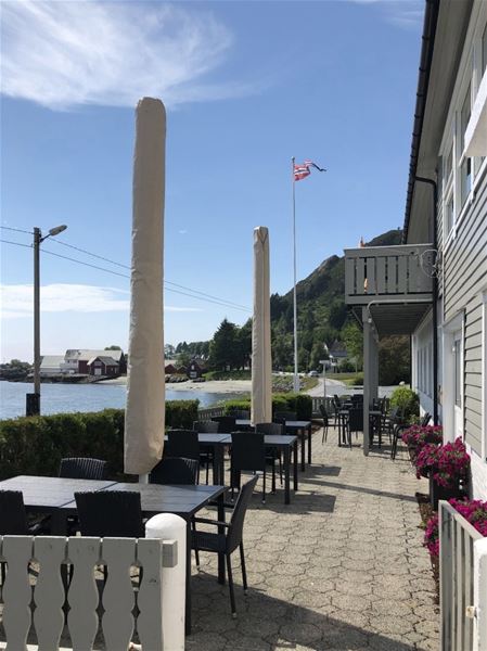 Outdoor cafe patio with tables, closed umbrellas, and a flag beside a waterfront road and hillside houses..