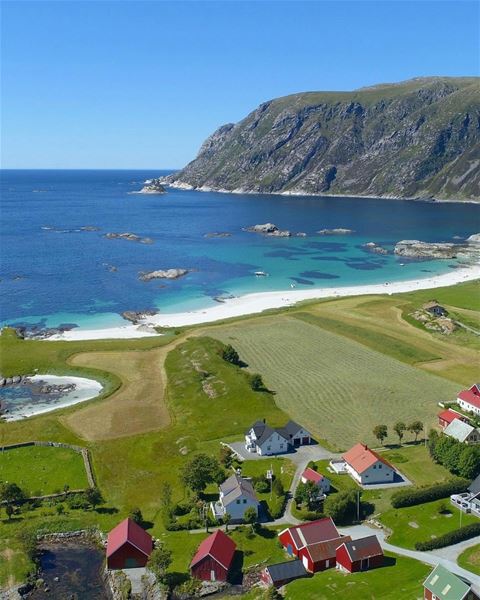 Aerial view of red-roof farm buildings by green fields and a white sand beach with turquoise sea and rocky cliffs..