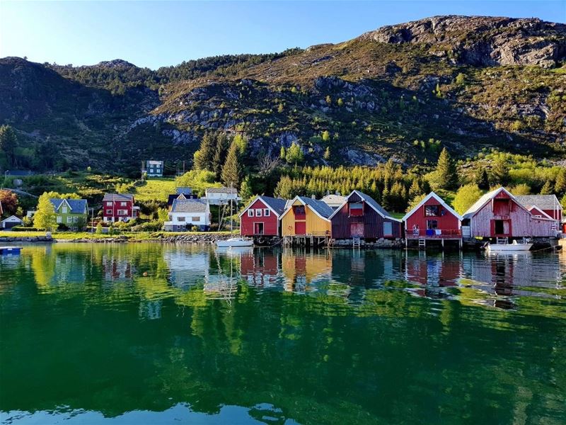 Red and yellow wooden boathouses line a calm fjord, reflecting in green water beneath rocky, tree-covered hills..