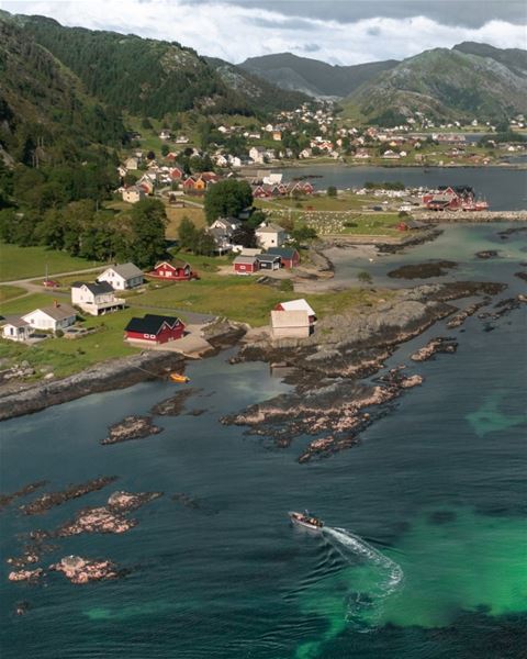 Aerial view of a coastal village with scattered houses, rocky shoreline, green hills, and a boat on turquoise water..