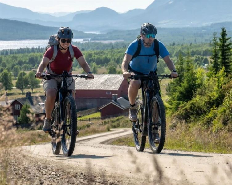Two people cycling on a gravel road in an open landscape with valley and mountains in the background.