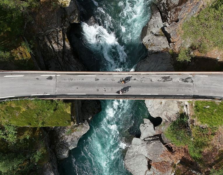 Aerial view of a bridge crossing a narrow river with rapids in mountainous terrain.