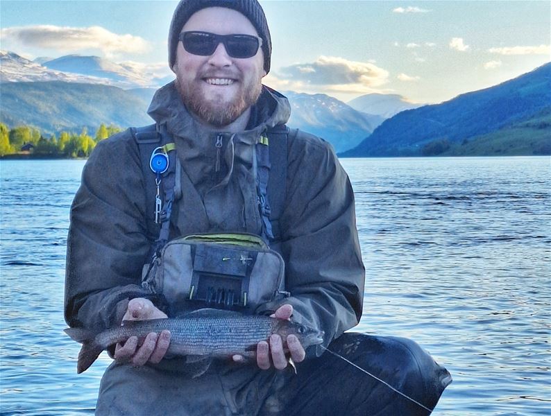 Person holding a fish by a lake, with mountains and water in the background.