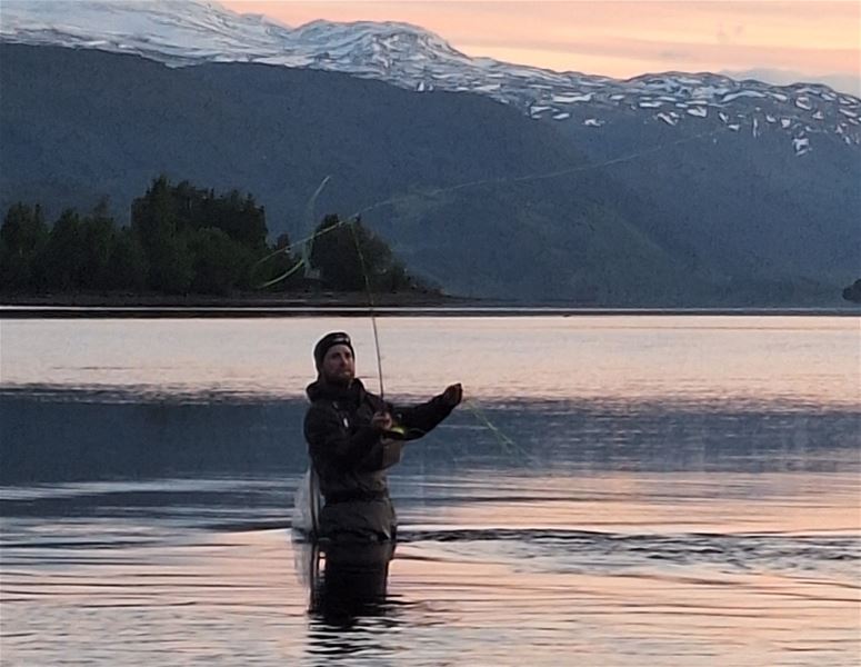 Person standing in the water fly fishing in a lake at dusk, with mountains in the background.