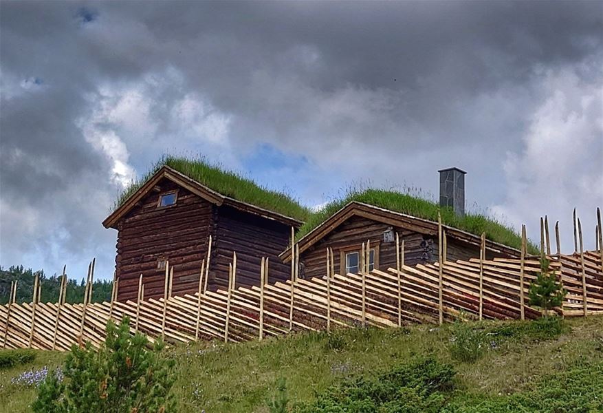 Traditionell seter med trebygninger og gjerder i fjellandskap under dramatisk himmel.