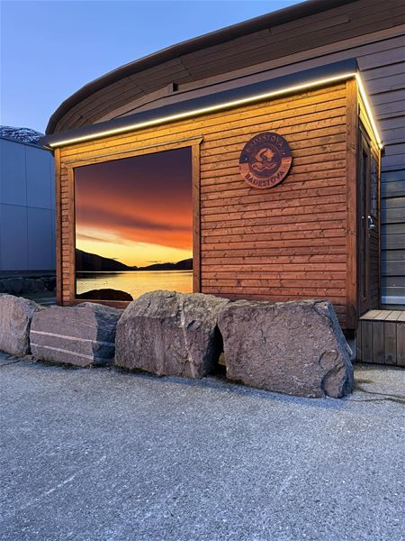 Small wooden building with LED trim and a large window reflecting a sunset over water, with boulders in front..