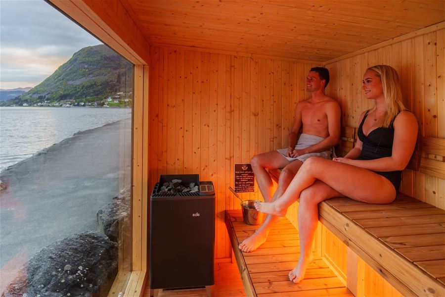 Two people sit on wooden benches in a sauna beside a large window showing a coastal road, sea, fjord and mountain..