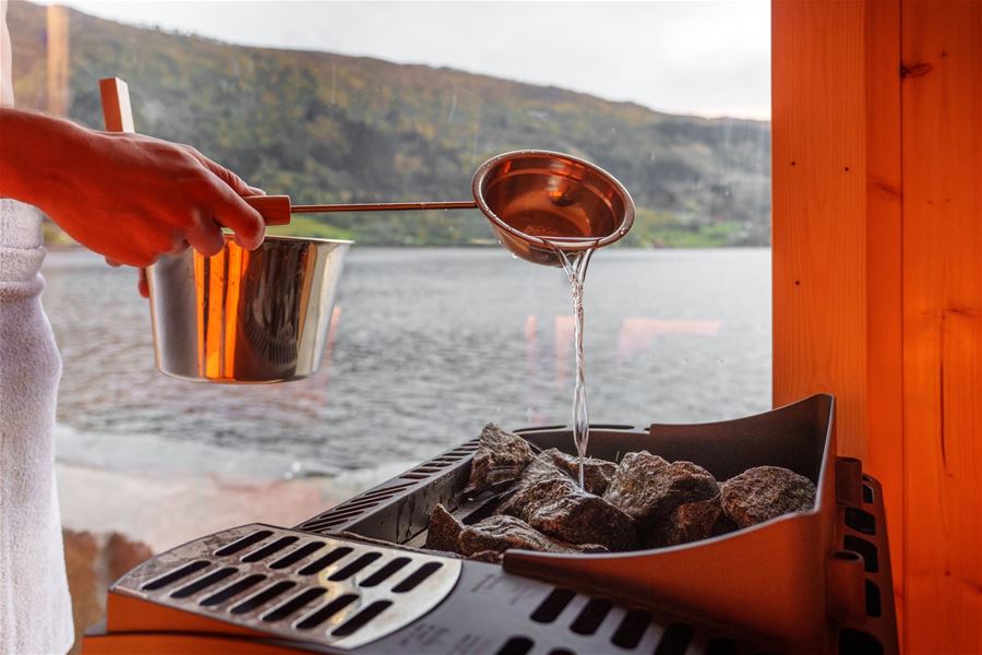 A hand pours water from a metal ladle onto hot sauna stones by a window overlooking a lake and hills in Nordfjordeid..