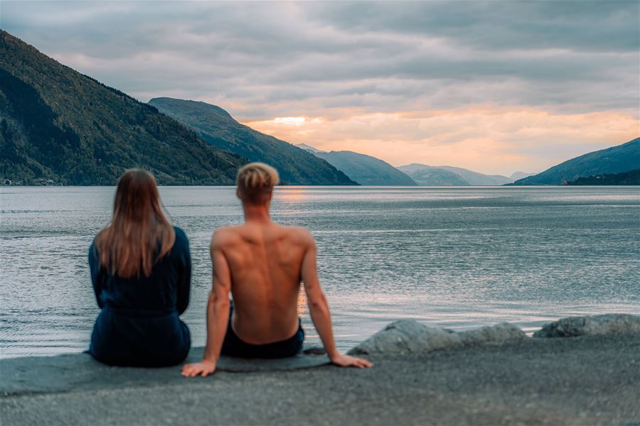Two people sit on a rocky shore facing a calm fjord and forested mountains under an overcast sunset sky at Nordfjordeid..