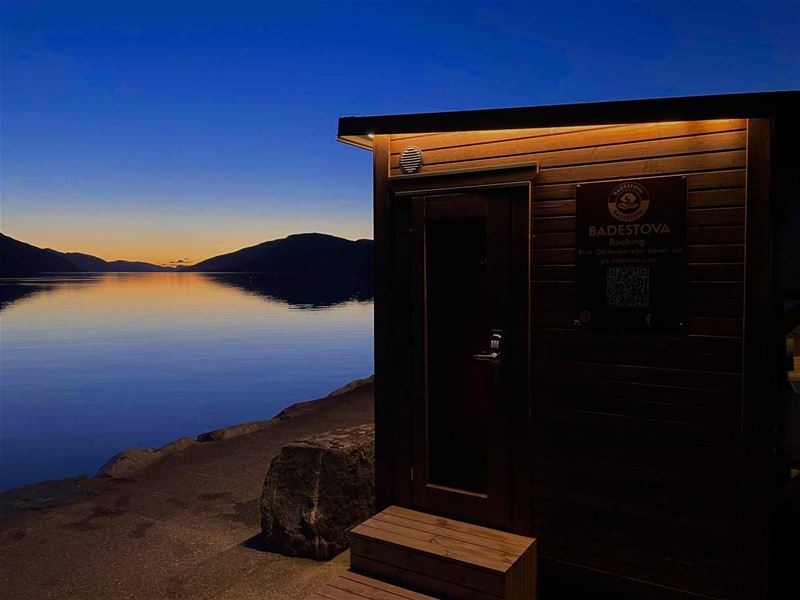 Badstova Sauna at Nordfjordeid with warm light by a calm fjord at dusk, with mountains silhouetted against a fading sky..