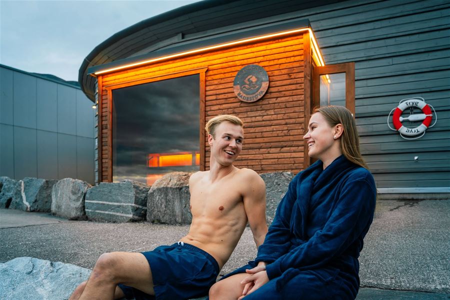 Two people sit on rocks outside a wooden sauna at Nordfjordeid by Nordfjorden building at dusk, with warm lights, a window, and a lifebuoy..