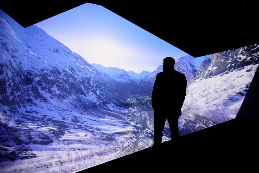 Ein person i silhuett står framfor ein stor projeksjon av snødekte fjell og dal under klar blå himmel..