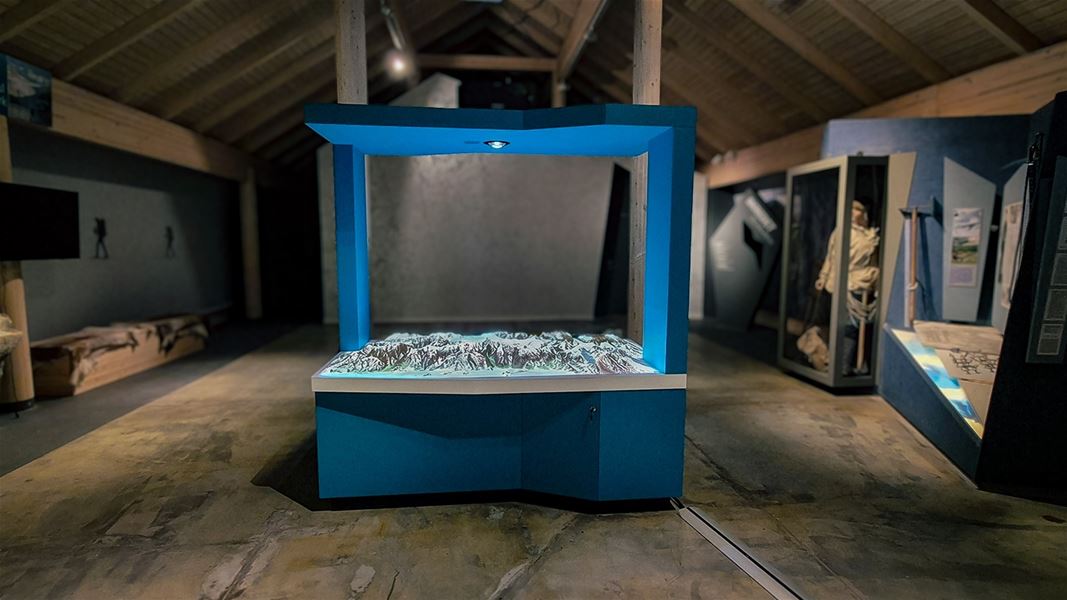 Innandørs utstilling med blå monter som lyser opp ein topografisk fjellmodell, omgitt av museumspanel og vitrinar..