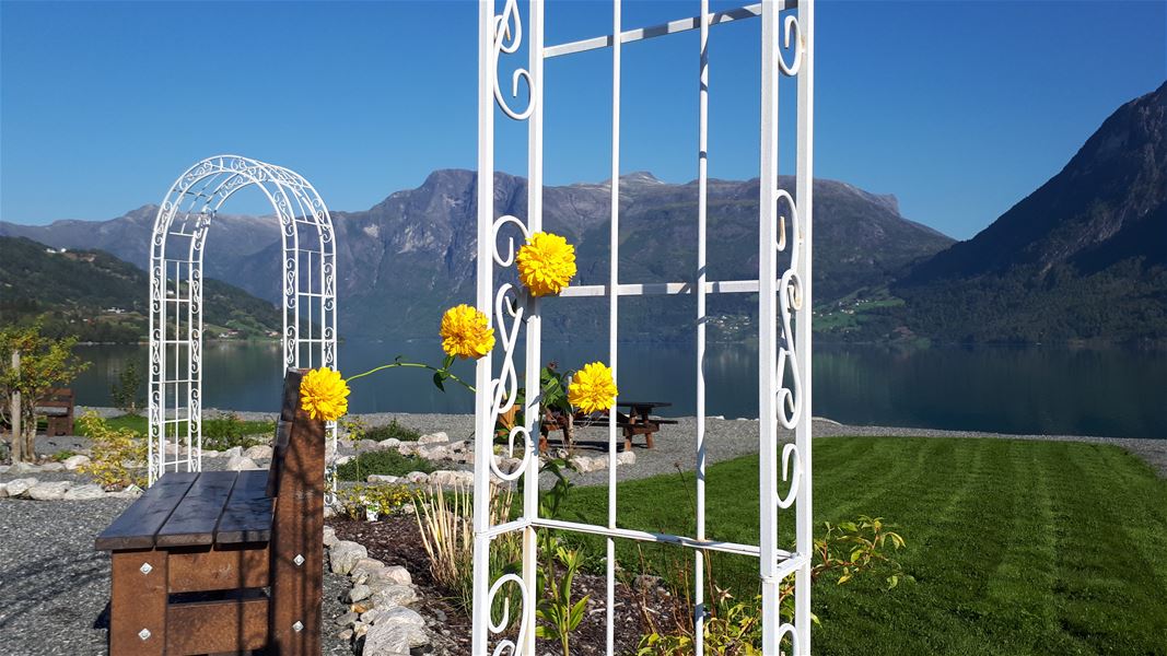 Kvit metallhageport og gule blomar i forgrunnen, med fjord, grøne bakkar og fjell under blå himmel..