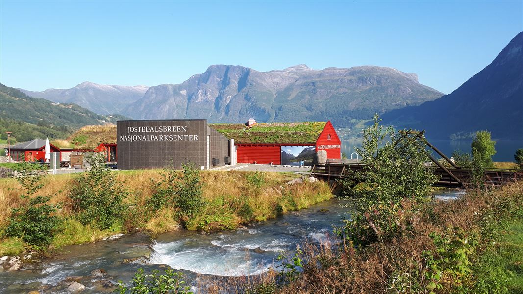 Raude bygg ved Jostedalsbreen nasjonalparksenter, med elv i forgrunnen og fjell og fjord under klar blå himmel..