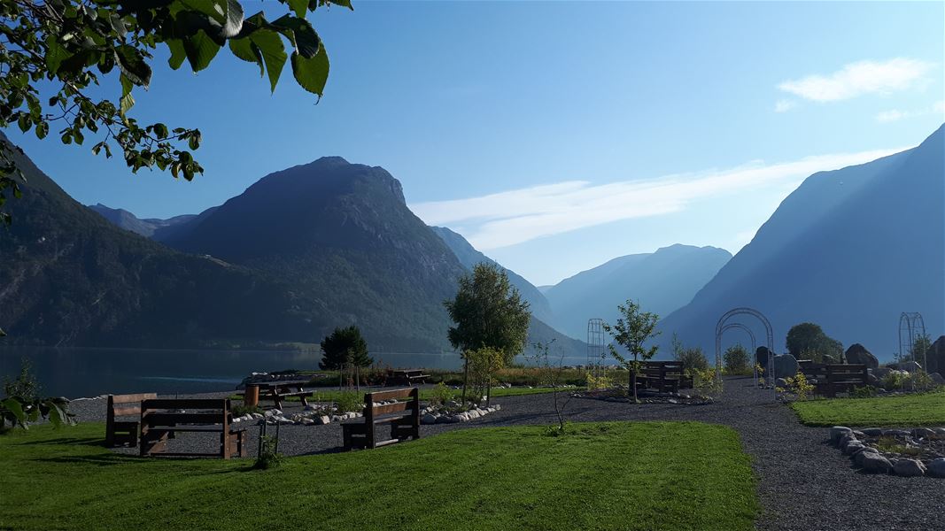 Grøn park med grusgangar og benker ved ein stille fjord, med store fjell og blå himmel i bakgrunnen..