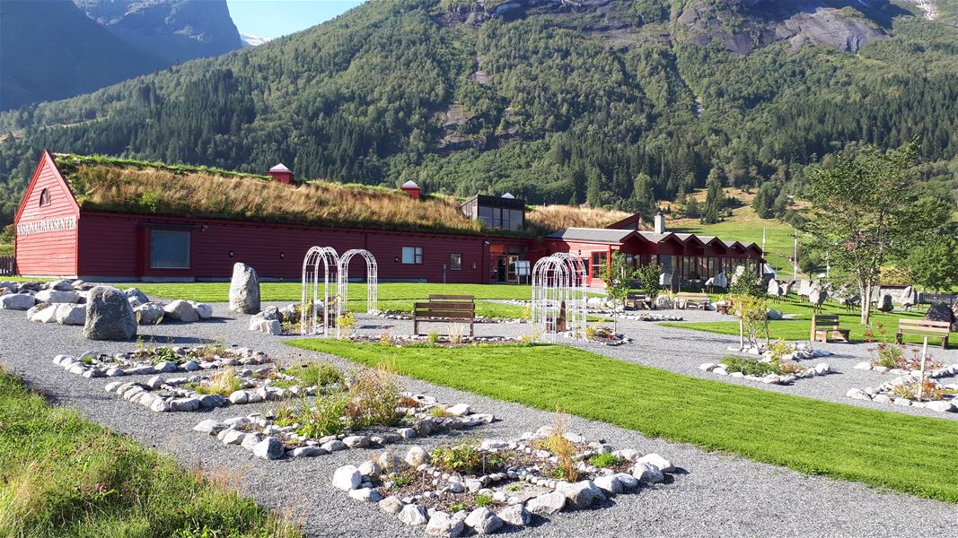 Raud bygning med torvtak ved grusganger, steinbed og benkar, med skogkledde fjell i bakgrunnen..