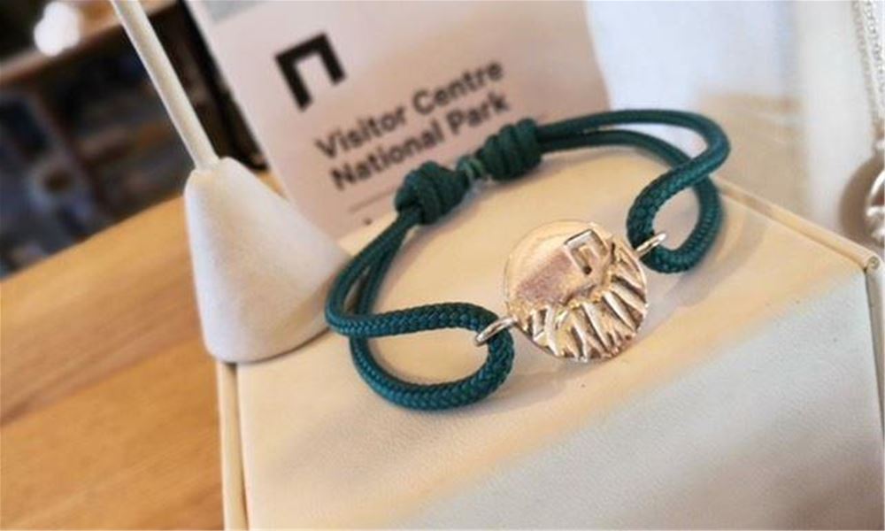 Nærbilete av grønt tauarmband med sølvfarga anheng, lagt på ei lys pute med skilt i bakgrunnen..