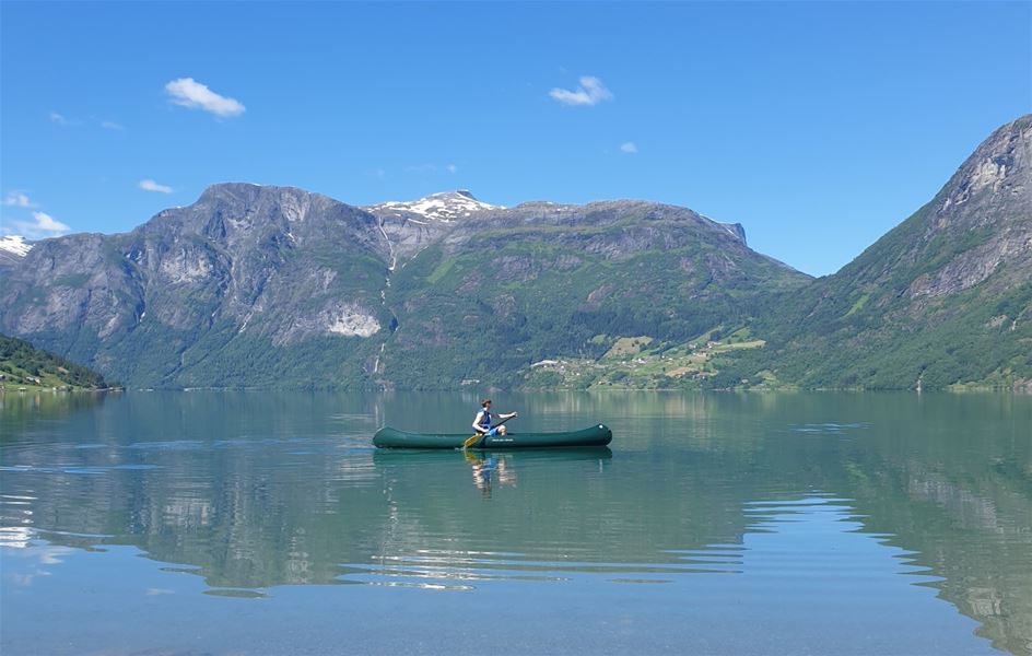 Ein person padlar ein grøn kano på spegelblankt vatn framfor grøne fjell med snøflekkar og blå himmel..