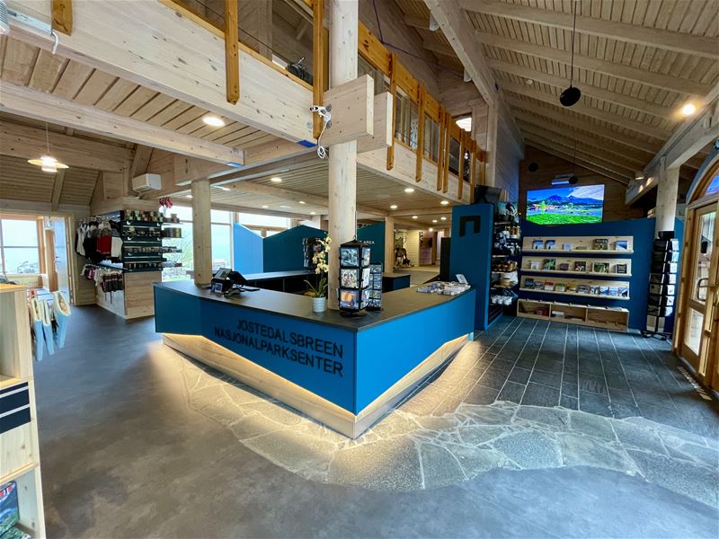 Innandørs besøkssenter i tre med blå resepsjonsdisk, suvenirhyller, brosjyrer, vindauge og skjerm på veggen..