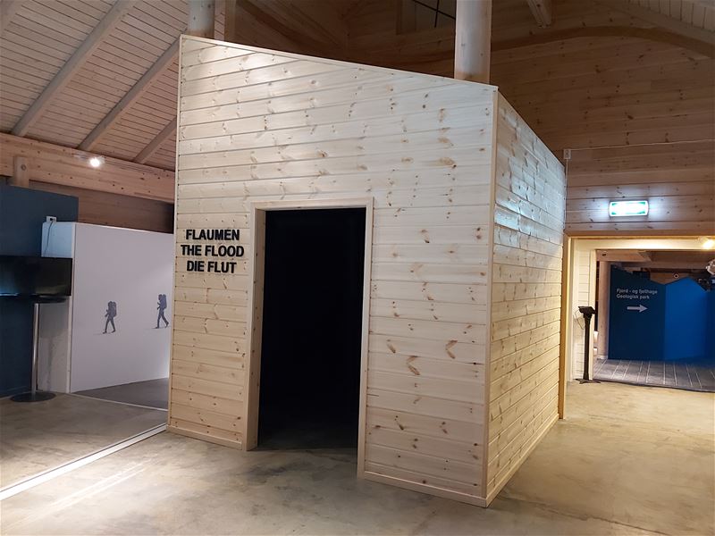 Innandørs utstilling med stor treboks, open dør og veggtekst «FLAUMEN / THE FLOOD / DIE FLUT» i eit trebygg..