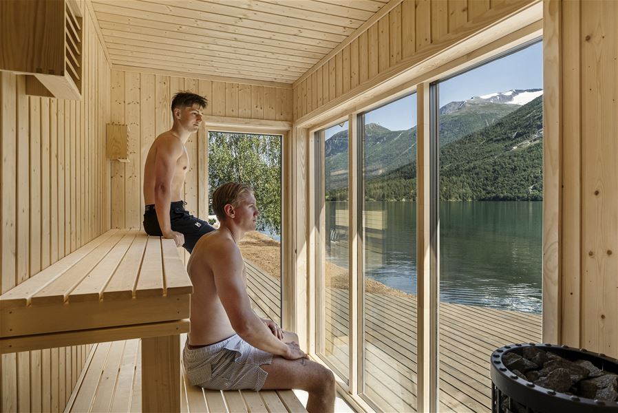 Two shirtless men sit on wooden benches inside a sauna, looking through large windows at a lake and mountains..