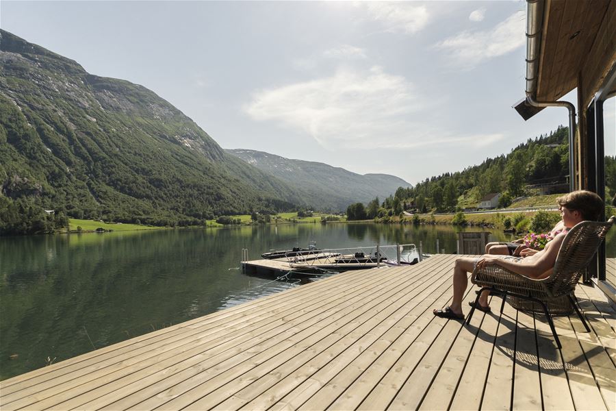 Two people sit in wicker chairs on a wooden deck overlooking a calm lake, docked boats, and green mountains..