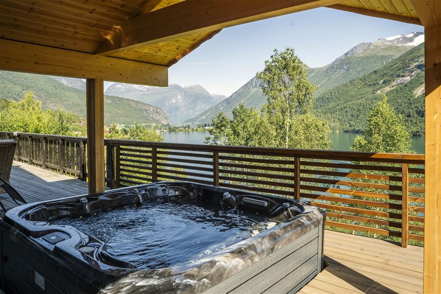 A bubbling hot tub on a wooden covered deck overlooks a calm lake, green trees, and distant mountains..