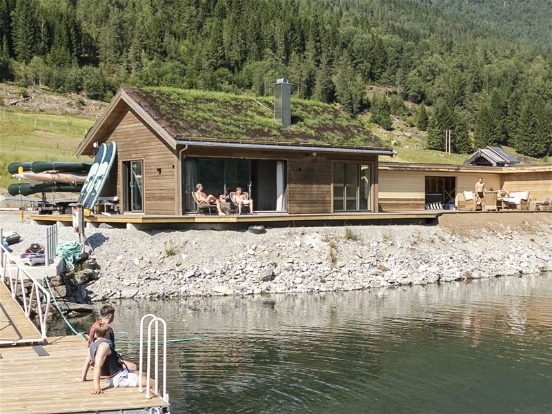 Wooden lakeside cabin with a green roof, people relaxing on a deck, kayaks nearby, and forested hills behind..