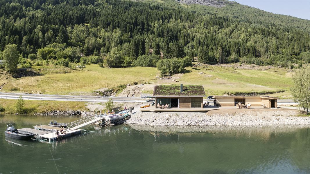 Modern wooden cabin with a green roof beside a calm lake, with a dock, boats, and forested hills behind..