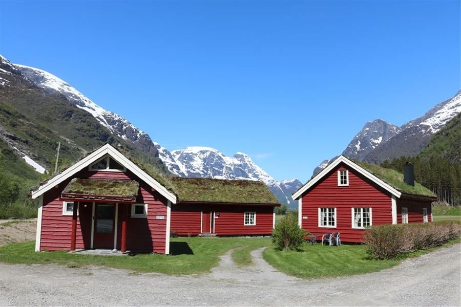 Two red wooden cabins with grass roofs sit on green grass, framed by snowy mountains under a clear blue sky at Aabrekk Gard Trollbu in Oldedalen valley..