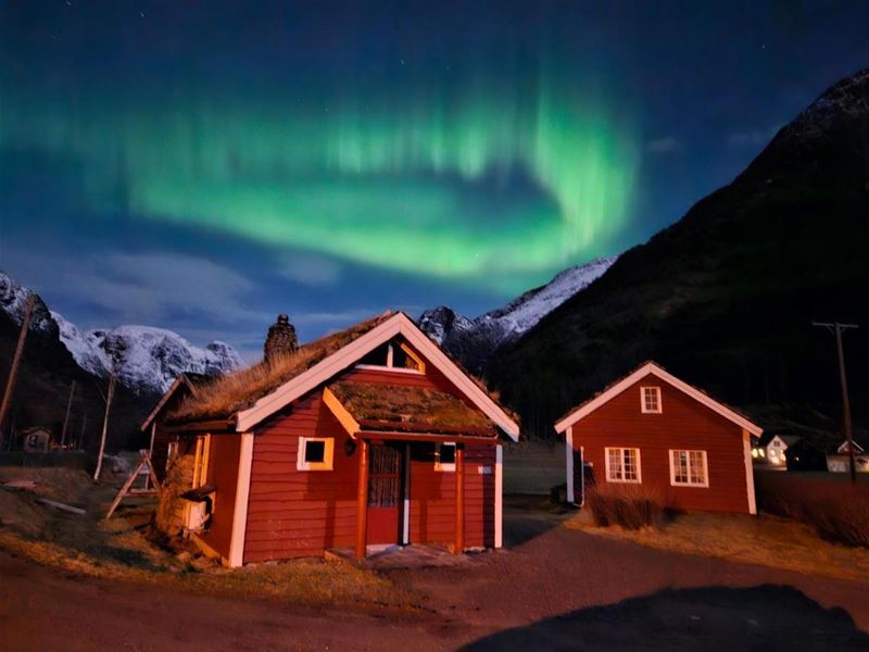 Green aurora swirls across a night sky above red wooden cabins, with snow-dusted mountains in the background at Aabrekk Gard Trollbu in Oldedalen valley..