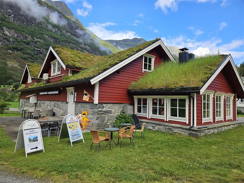 Red wooden cottage with a grass-covered roof, outdoor café tables and signs, set below green mountains under blue sky at Aabrekk Gard Trollbu in Oldedalen valley..