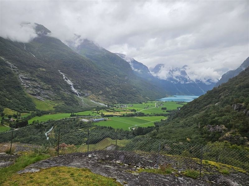 Wide view of a green valley between mountains, with waterfalls, farmland, and a turquoise fjord under clouds..