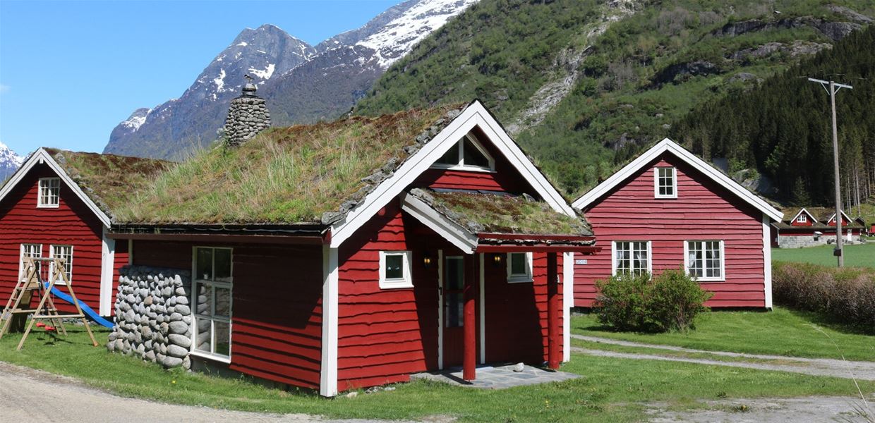 Two red wooden cabins with grass-covered roofs sit on green lawn, with forested and snowy mountains behind at Aabrekk Gard Trollbu in Oldedalen valley..