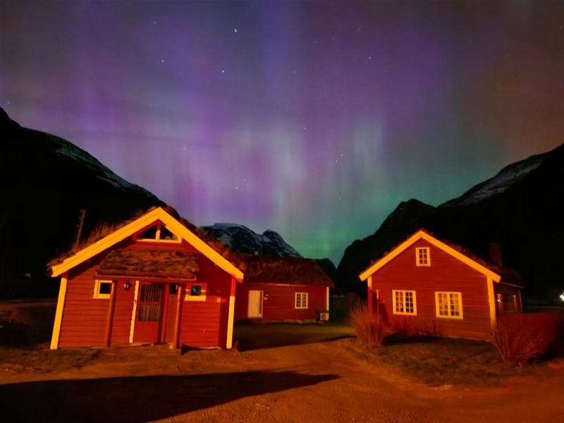 Two red cabins lit in warm light beneath a purple and green aurora sky, framed by dark mountains at night at Aabrekk Gard Trollbu in Oldedalen valley..
