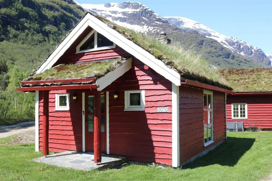 Red wooden cabin with white trim and grass-covered roof on a green lawn, with snowy mountains behind, at Aabrekk Gard Trollbu in Oldedalen valley.