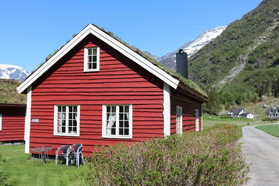 Red wooden cabin with white-trim windows and grass roof beside a lawn, road, and snowy mountains under blue sky at Aabrekk Gard Trollbu in Oldedalen valley.