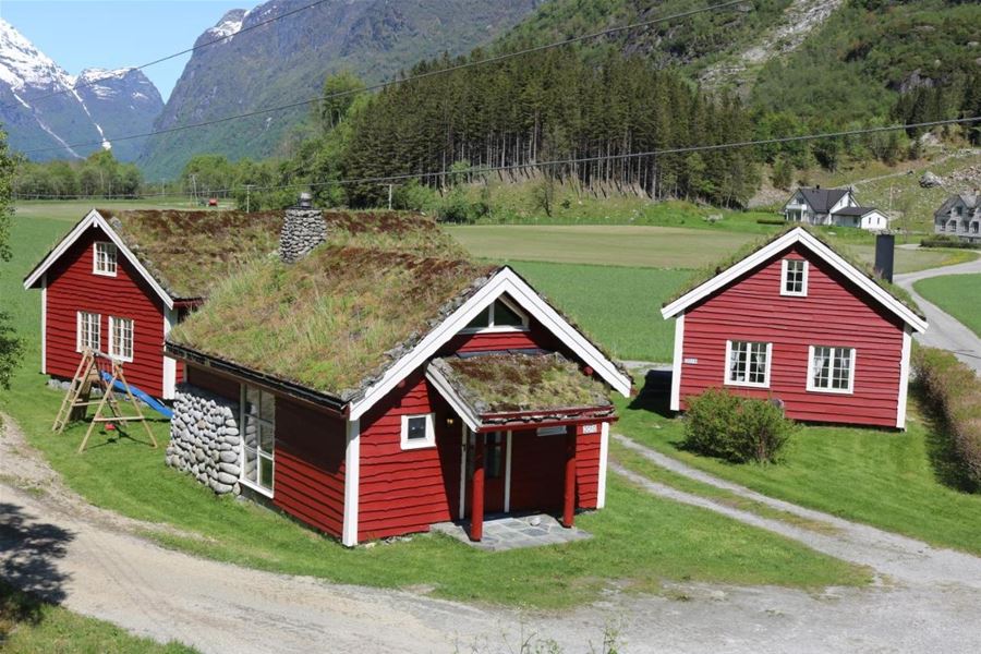 Three red wooden houses with grass roofs sit on green fields, with forested hills and snowy mountains behind, at Aabrekk Gard Trollbu in Oldedalen valley.