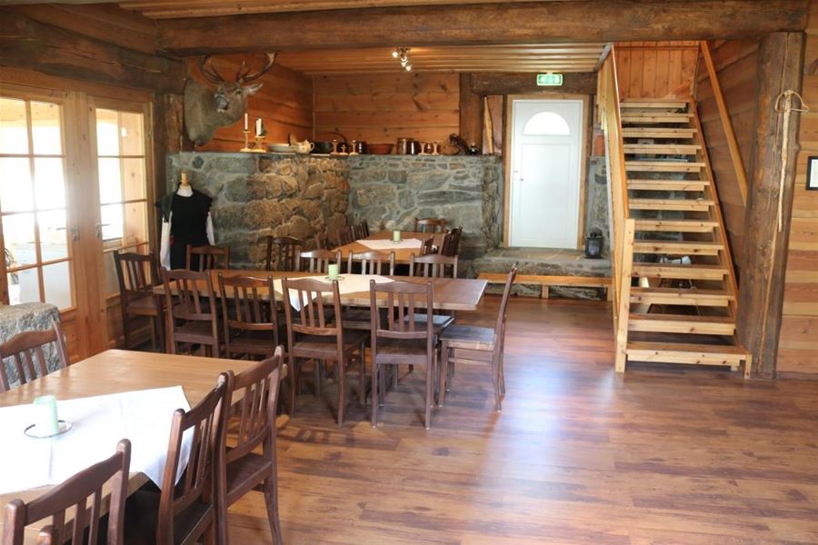 Wood-paneled lodge dining room with long tables and chairs, a stone wall with a mounted deer, and stairs by a white door, at Aabrekk Gard Trollbu in Oldedalen valley.