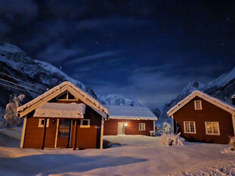 Three snow-covered wooden cabins lit by warm lights sit in a winter landscape beneath a dark, starry sky, at Aabrekk Gard Trollbu in Oldedalen valley.