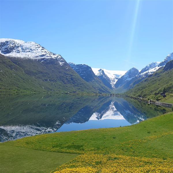 Snødekte fjell kring ein stille fjord som speglar landskapet, med grøne bakkar og blå himmel i Oldedalen.
