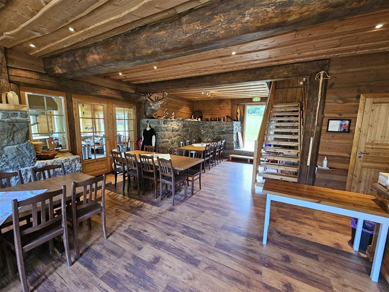 Innvendig tømmerhytte med trebord og stolar, steinvegg og trapp, samt open dør mot grønt uteområde på Aabrekk Gard - Kafé og Restaurant i Oldedalen.