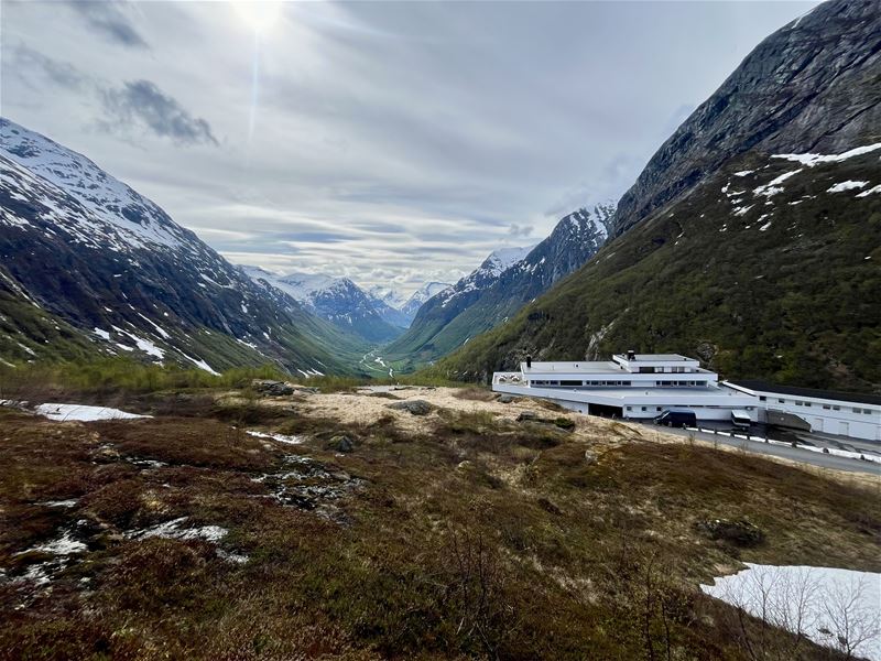 Utsyn over ei grøn fjelldal med snøflekkar på fjellsidene, skydekke og eit bygg nede til høgre..