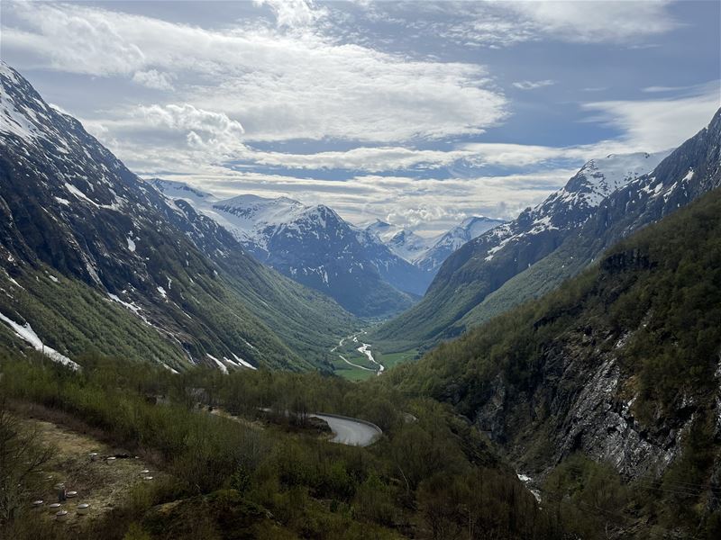 Vid utsikt over ein grøn dal mellom snødekte fjell, med svingete veg og elv under ei skydekka blå himmel..