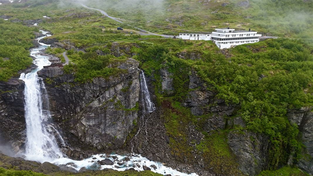 Luftfoto av ein kraftig foss som stuper ned i ein steinrøys, med grøne fjellsider og eit kvitt hotell på kanten..