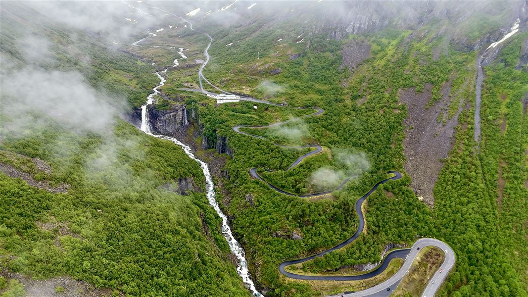 Luftfoto av svingete fjellveg gjennom grønt dalføre med foss og elv, omkransa av skyer og bratte fjellsider..
