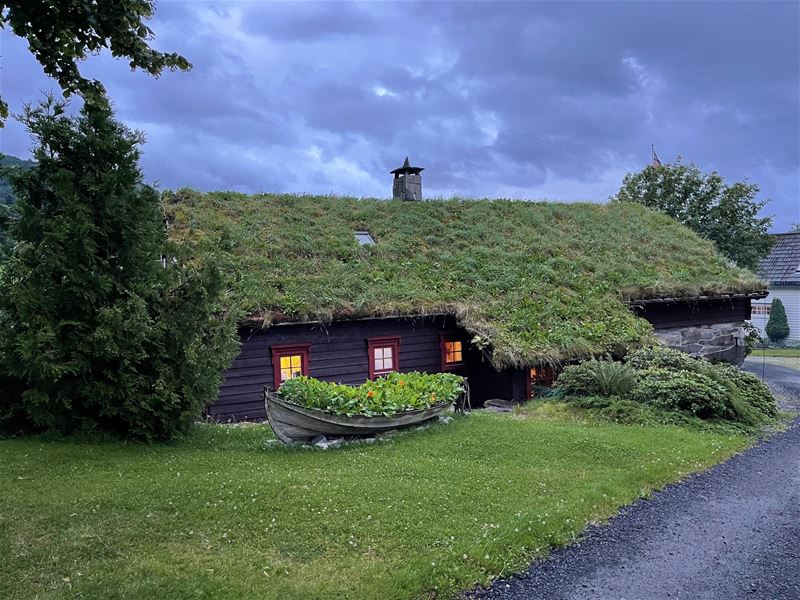 Orheimstunet i Nordfjord - Mørkt trehus med torvtak og raude vindauge, båtblomsterbed på grasplen under dramatisk skydekke..